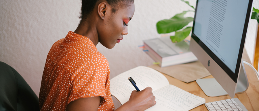A person in a polka dot shirt writes in a notebook at a desk with a computer. The setting includes books and a plant, suggesting a study or work environment.